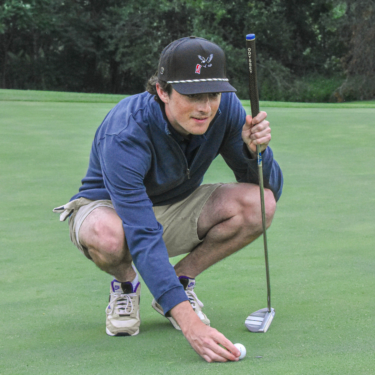 A man crouches on a golf green holding a putter, wearing a navy blue long-sleeve shirt, khaki shorts, and a black baseball cap with a white logo.