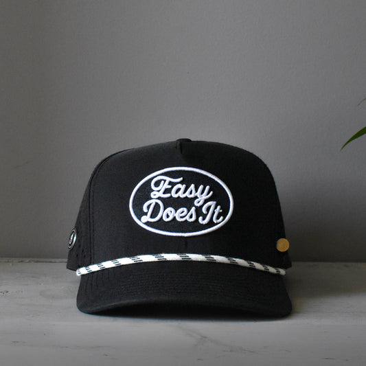 A black baseball cap with a white embroidered oval logo reading 'Easy Does It' on the front, a white-and-black patterned horizontal stripe across the brim, and a small gold button on the side, photographed against a plain gray wall.