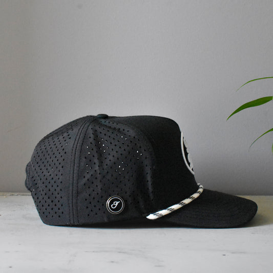A black mesh baseball cap with a white and black patterned rope detail on the brim, resting on a light marble surface against a gray wall with a green plant leaf in the background.