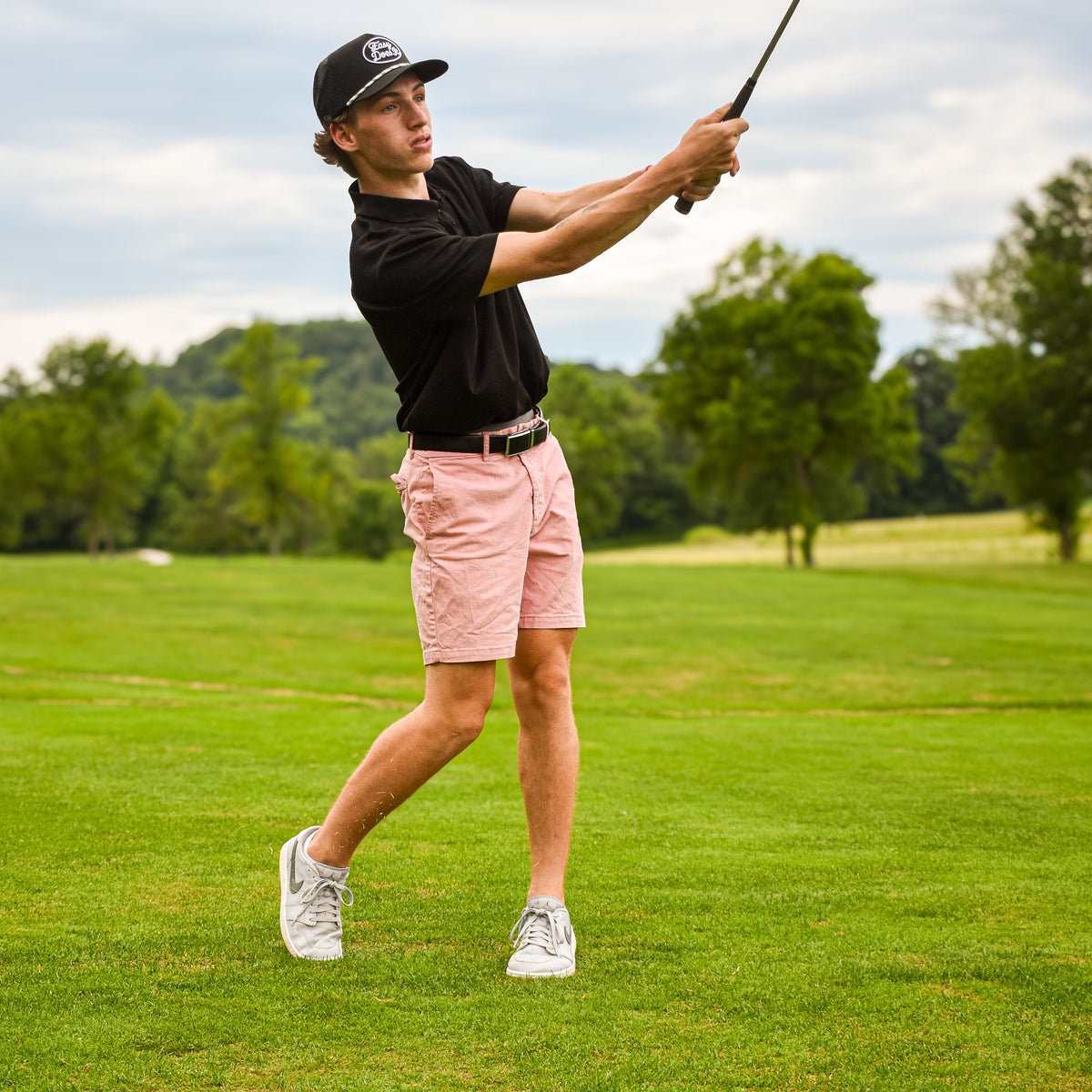 A man in a black polo shirt, pink shorts, and a black baseball cap is captured mid-swing on a green golf course.