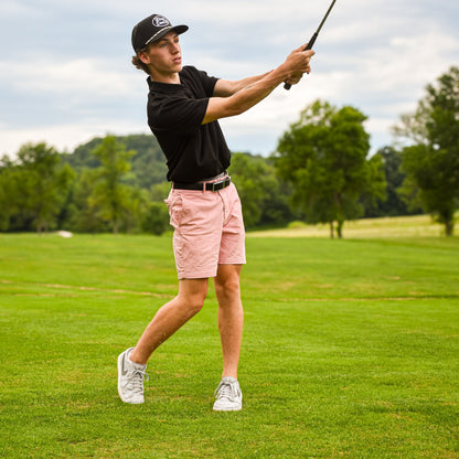 A man in a black polo shirt, pink shorts, and a black baseball cap is captured mid-swing on a green golf course.