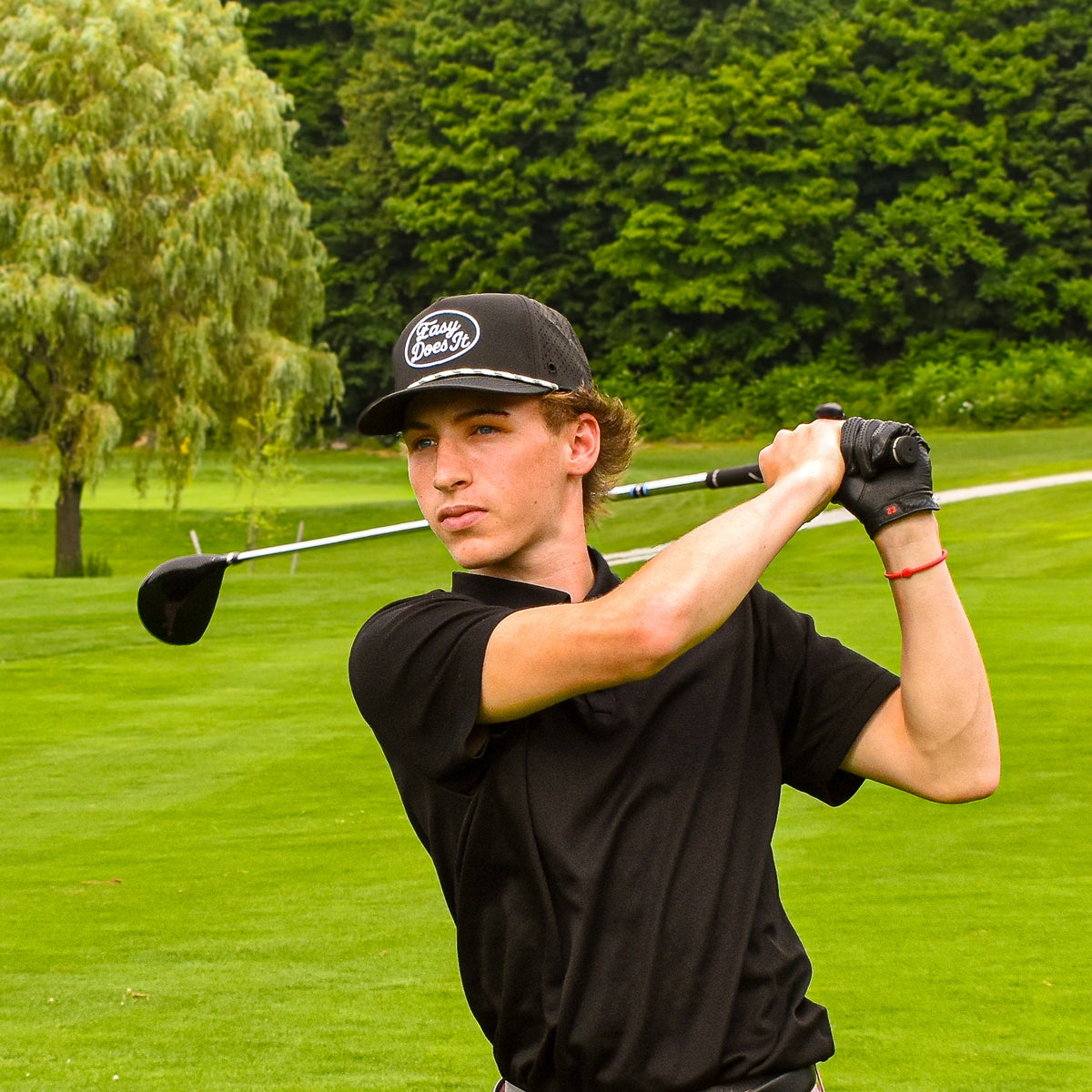 A man in a black polo shirt, pink shorts, and a black cap is captured mid-swing on a lush green golf course.
