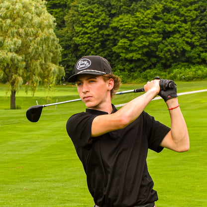 A man in a black polo shirt, pink shorts, and a black cap is captured mid-swing on a lush green golf course.