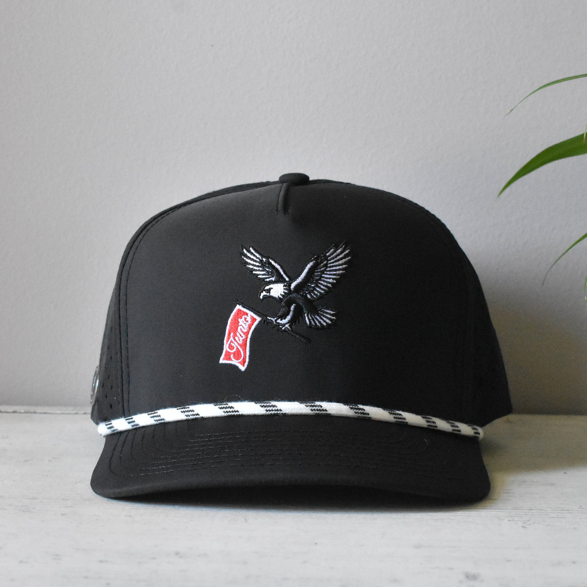 A black snapback cap featuring an embroidered eagle holding a red and white 'Junto' flag, with a white and black checkered stripe detail along the brim, displayed on a light surface against a neutral gray background.