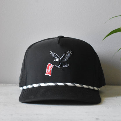 A black snapback cap featuring an embroidered eagle holding a red and white 'Junto' flag, with a white and black checkered stripe detail along the brim, displayed on a light surface against a neutral gray background.