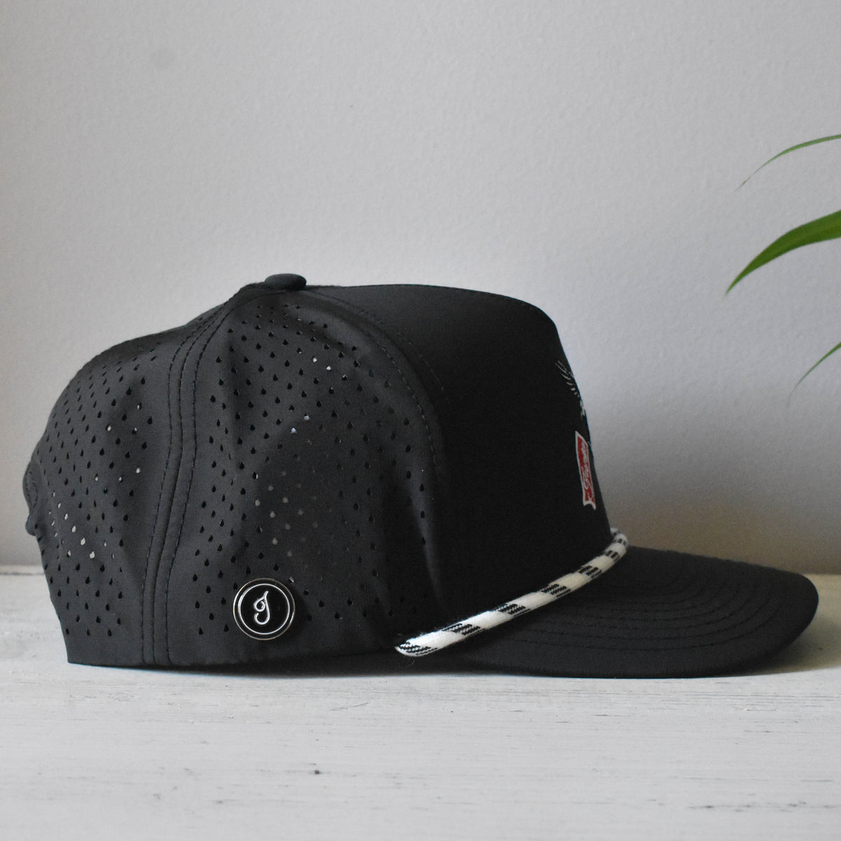 A black perforated snapback cap with a white and black patterned rope detail on the brim, resting on a light wooden surface against a neutral gray wall.