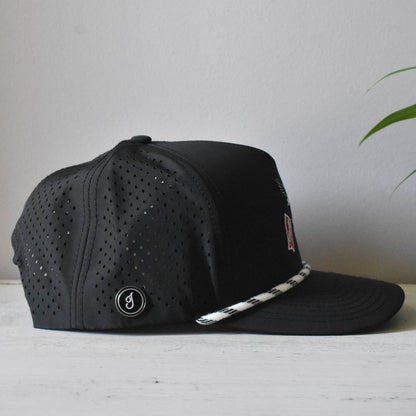 A black perforated snapback cap with a white and black patterned rope detail on the brim, resting on a light wooden surface against a neutral gray wall.