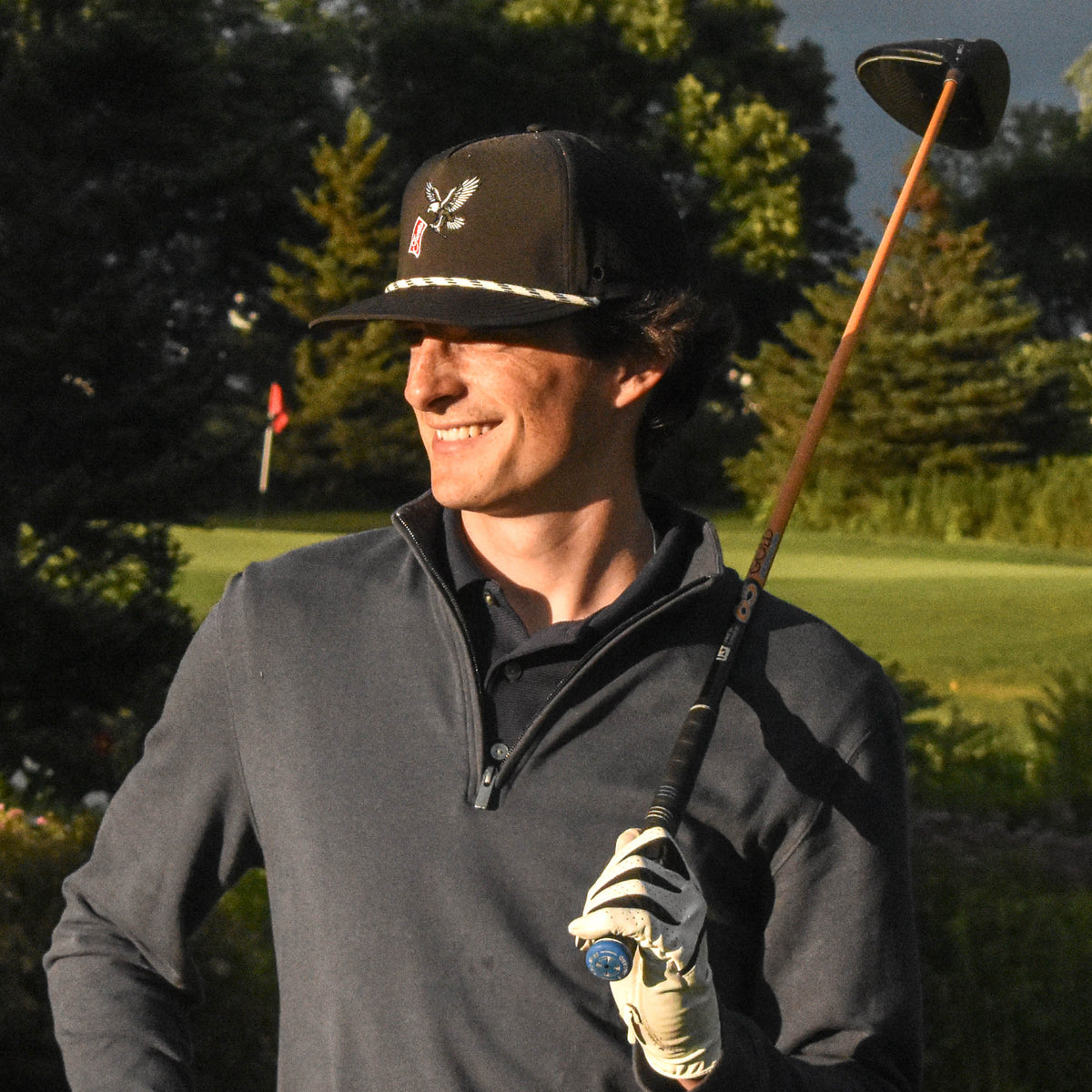 A man wearing a dark baseball cap, gray quarter-zip pullover, and golf glove stands on a golf course holding a golf club under a dramatic cloudy sky.