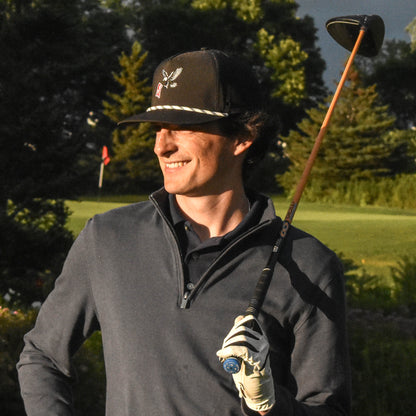 A man wearing a dark baseball cap, gray quarter-zip pullover, and golf glove stands on a golf course holding a golf club under a dramatic cloudy sky.