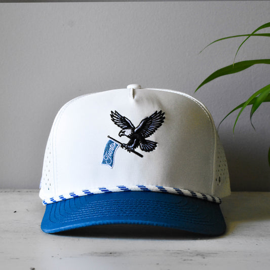 A white and blue trucker-style cap with a black embroidered eagle holding a blue flag that reads 'Junto' displayed on a light surface against a gray wall with a green plant leaf in the background.