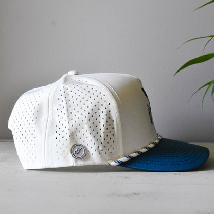 A white perforated baseball cap with a blue visor and a small circular logo badge, resting on a light gray surface against a gray wall with a plant leaf in the background.