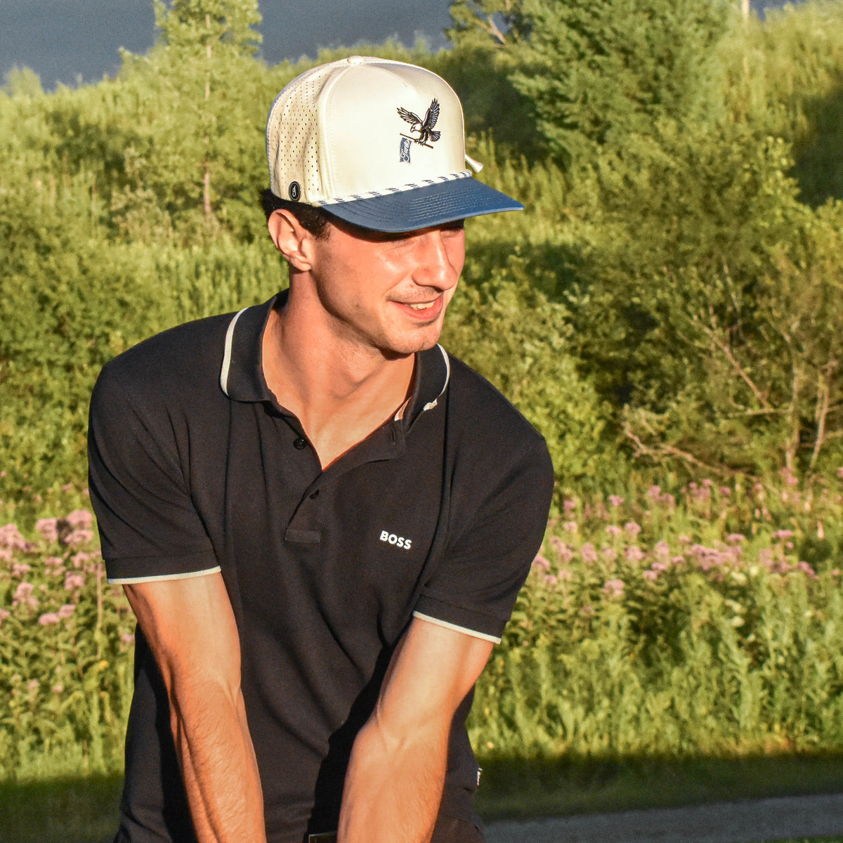 A man wearing a black polo shirt and a cream and navy baseball cap stands outdoors on a golf course or park with trees and a cloudy sky in the background.