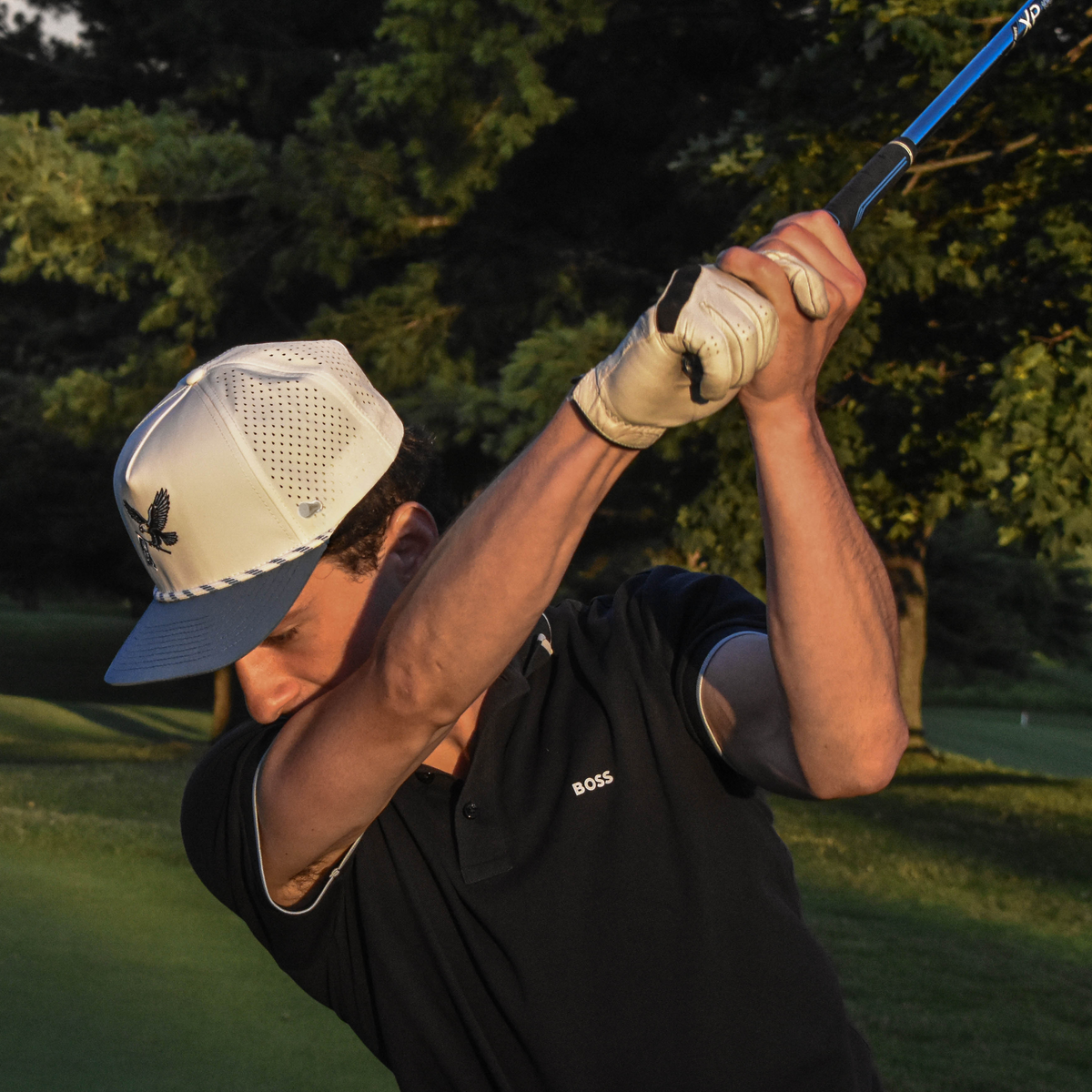 A golfer in a black polo shirt and white cap mid-swing on a golf course, with trees and grass in the background.