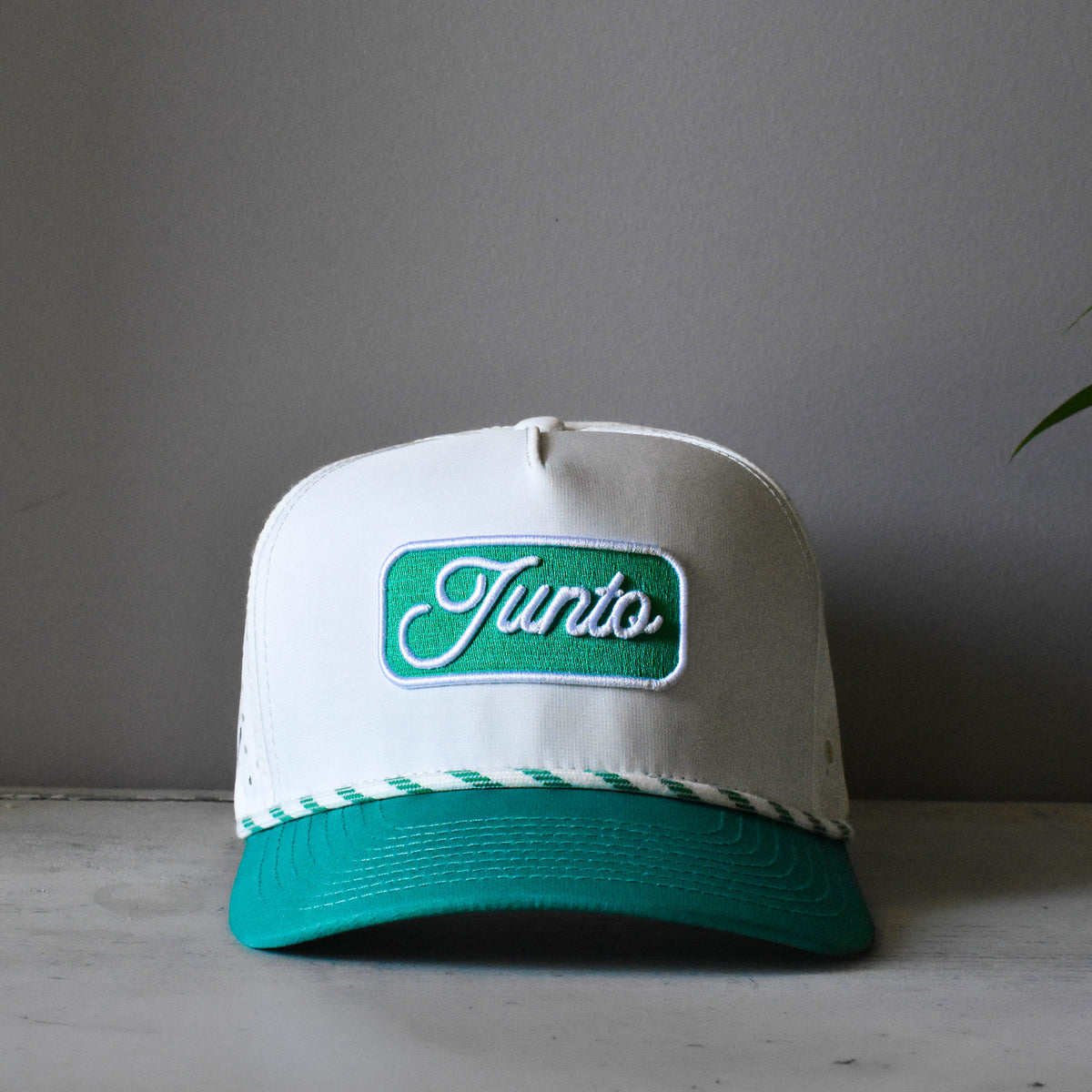 A white and teal trucker-style cap with a green embroidered patch on the front that reads 'Junto' in white script. The cap is resting on a light gray surface against a neutral gray background.