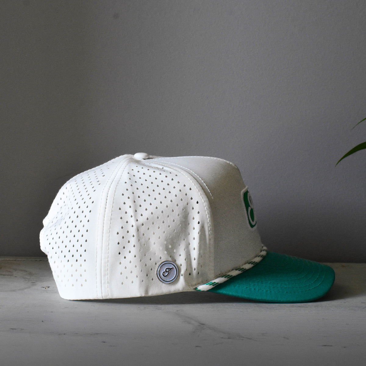 A white perforated baseball cap with a teal visor and a green magnetic ball marker on the side, resting on a light gray marble surface against a dark gray background.