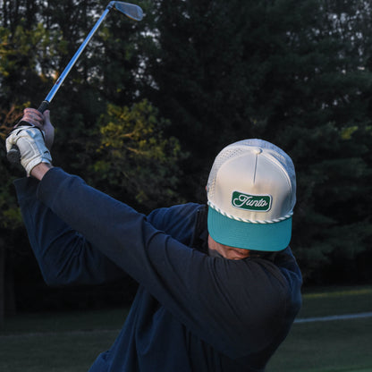 A person on a golf course wearing a white and teal cap with a green and white 'Junto' logo, a navy long-sleeve shirt, and a white glove, captured mid-swing with a golf club.