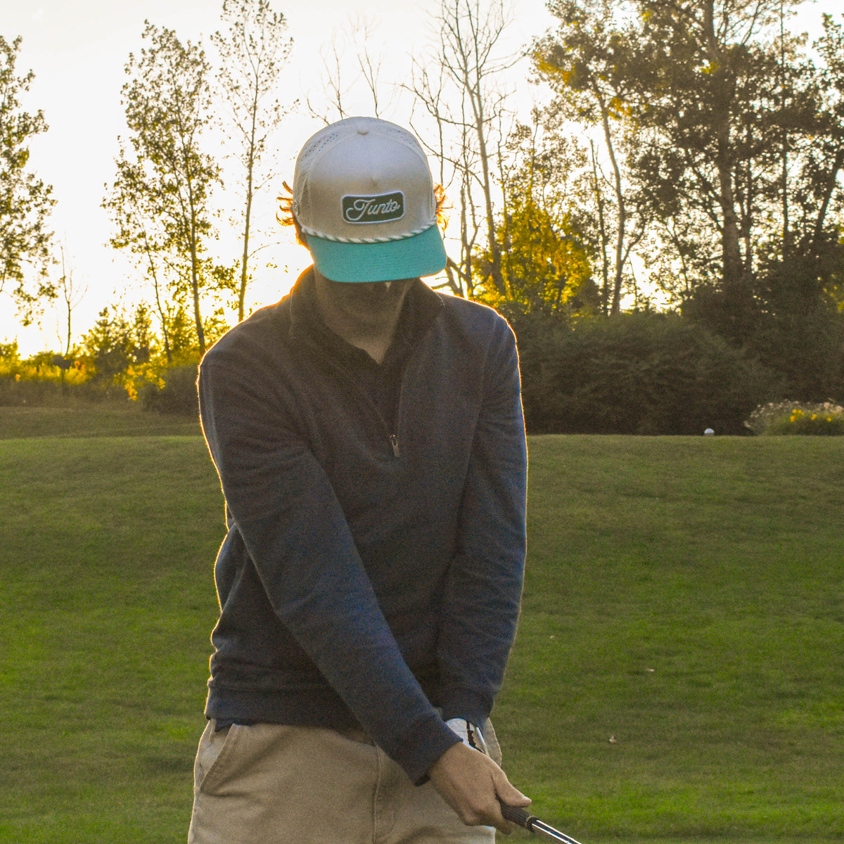 A man on a golf course wearing a white and teal cap, a dark long-sleeve shirt, and khaki pants, captured in warm golden hour light with trees in the background.